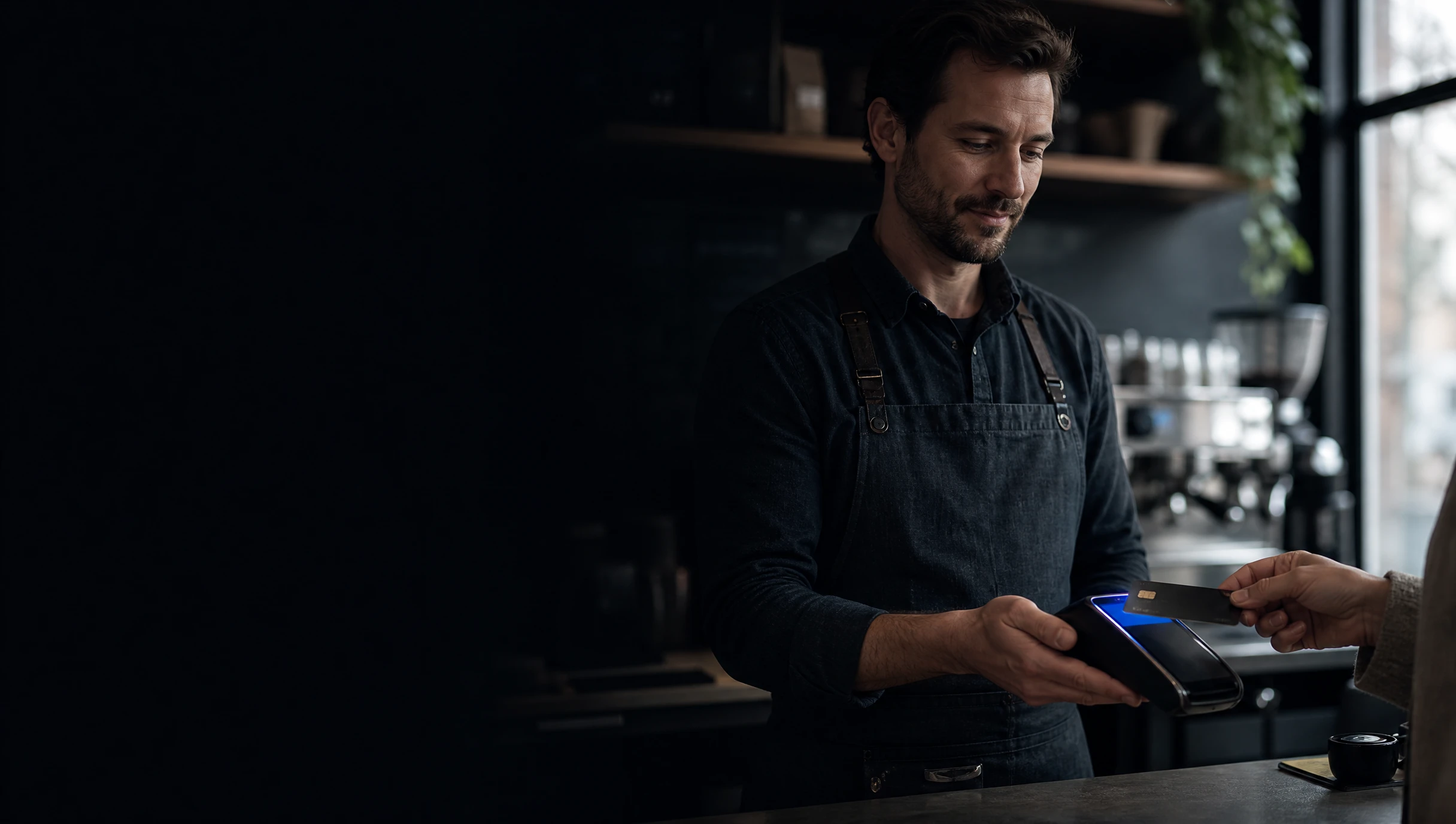 Café owner in an apron holding out a payment terminal as a customer taps a bank card at the counter